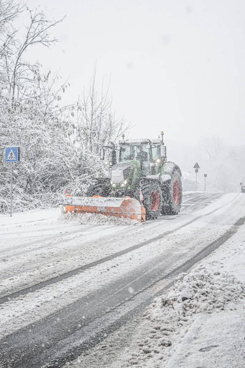 Traktor mit orangem Schneepflug räumt bei Schneefall eine Straße